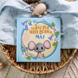 The book cover for Day You Were Born In May. A playful, illustrated mouse sat down surrounded by bluebells and lilies.